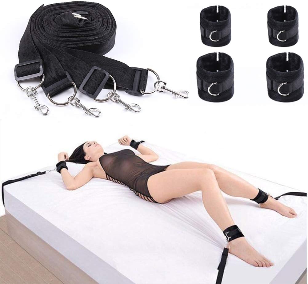 The image shows a person lying on a white bed with black under bed restraints attached to their wrists and ankles. The restraints appear to be made of nylon with metal rings, and there is a rolled-up black restraint lying beside the person.